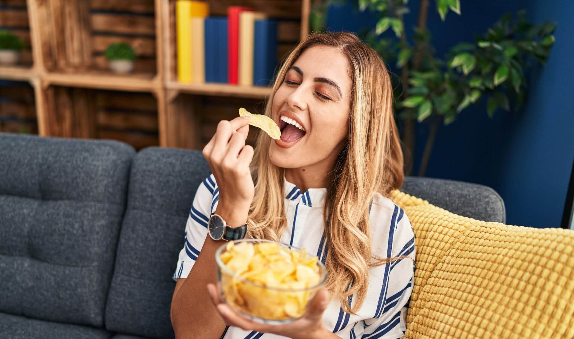 Woman sitting on a couch, smiling and eating potato chips improved by salt reduction.