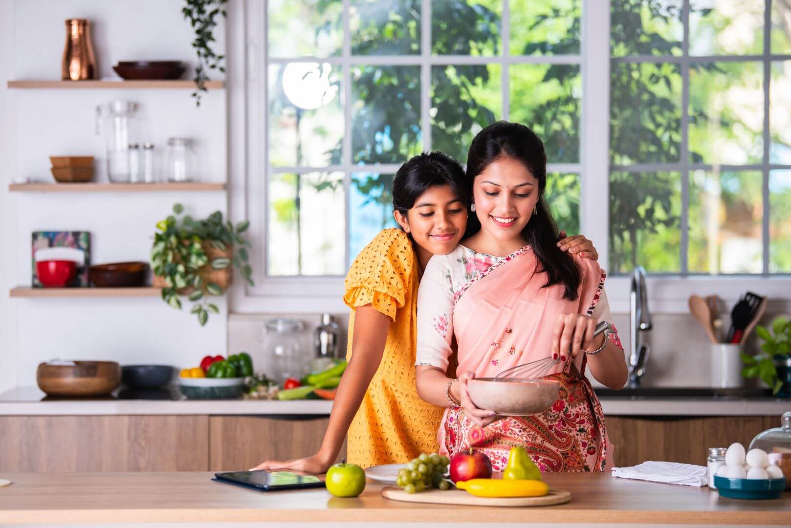 Mom and Generation Alpha daughter in India, hugging each other, smiling and mom cooking in the kitchen while daughter watches