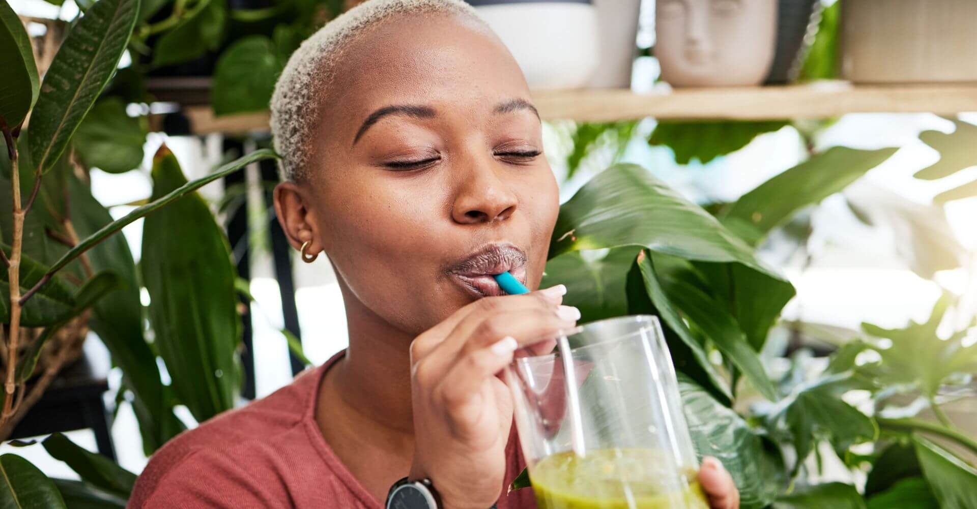 Woman with short hair and red shirt sipping green smoothie from a straw, with eyes closed and plants in background