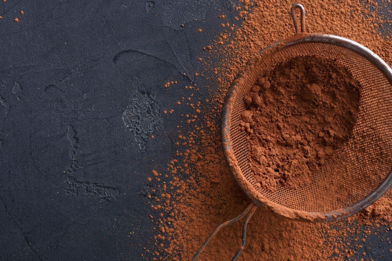 Cocoa powder in and around a large sieve, sitting on a black granite counter