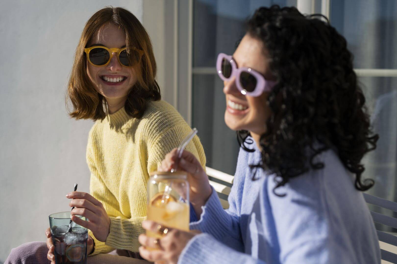 Two women (one with yellow sweater and sunglasses; other with purple sweater and sunglasses), sitting outside, smiling and drinking different iced beverages from straws