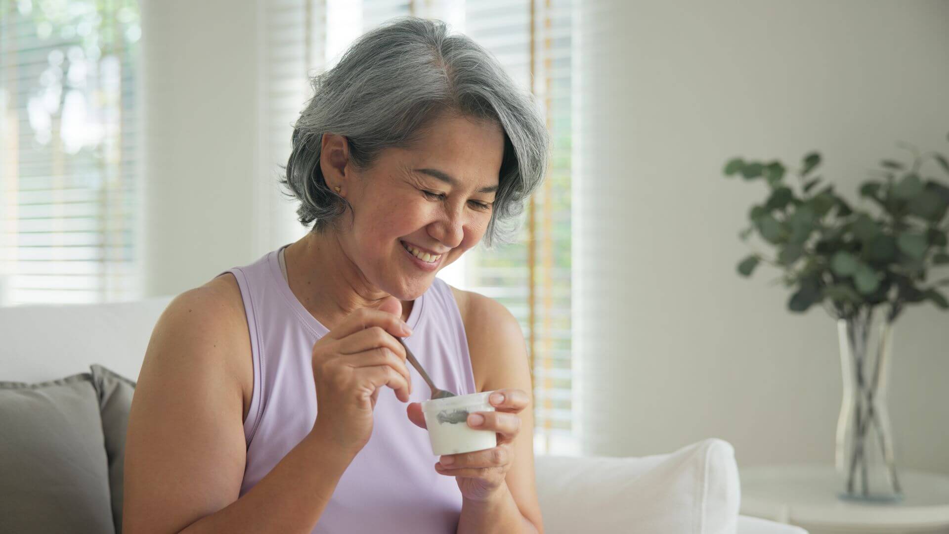 Senior woman sitting on couch, smiling and eating a yogurt snack that's been modulated with IFF FLAVORFIT [masking]
