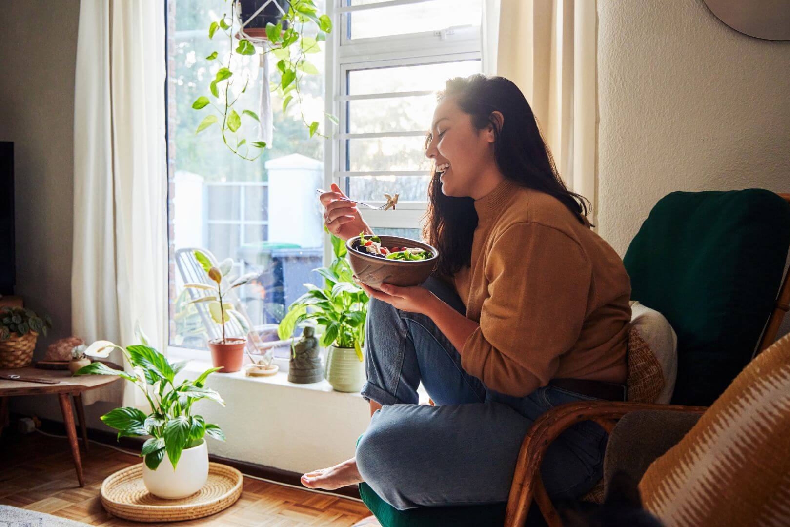 Woman who is a GLP-1 consumer eating a salad and sitting in a chair with leg up. Smiling and surrounded by plants in her apartment.