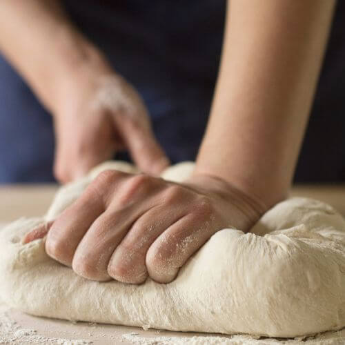 Mãos pressionando e sovando uma massa de pão sobre uma superfície enfarinhada, destacando a textura elástica da massa.