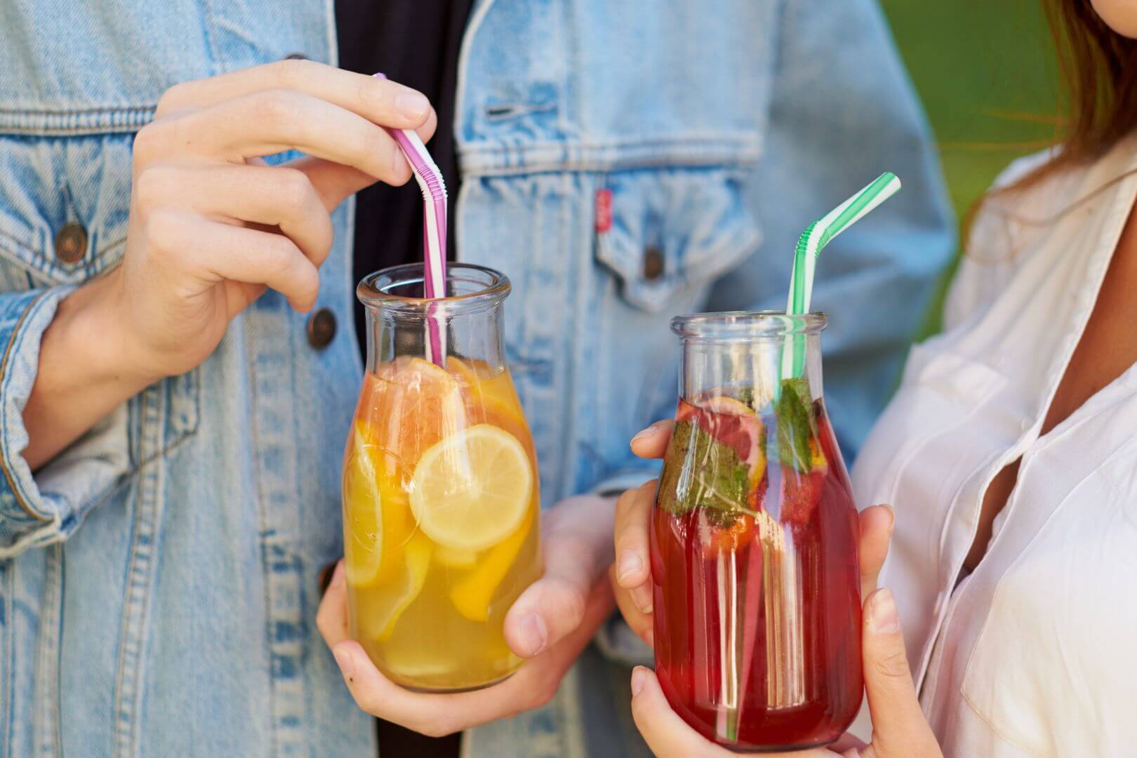 A man and a woman holding citrus-based beverages that are in glass bottles with straws (one is a red juice and one is an orange juice). Faces off camera