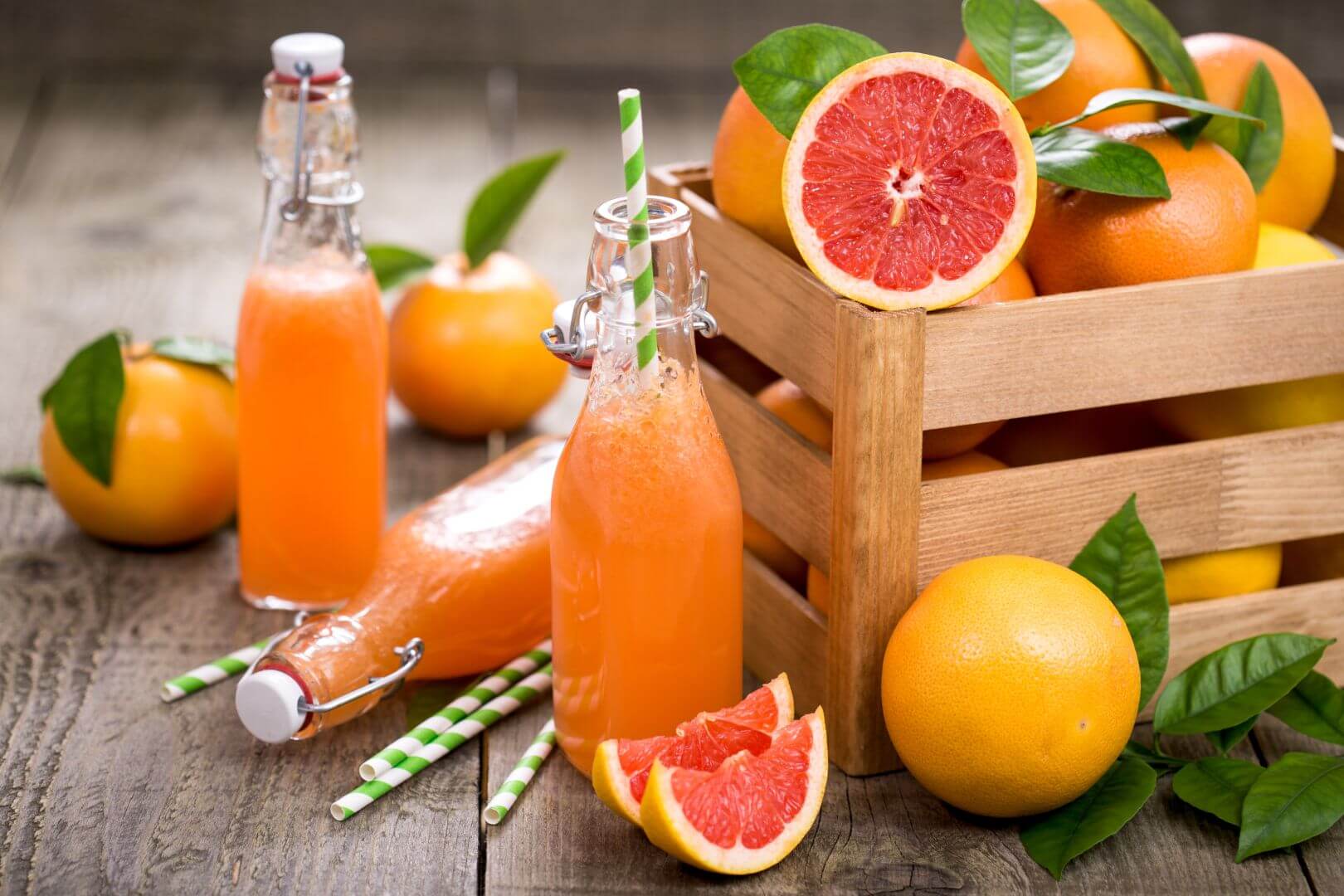 A crate of grapefruits (both whole and halved), sitting on a table next to bottles of grapefruit juice and straws, as well as more sliced and whole grapefruits.