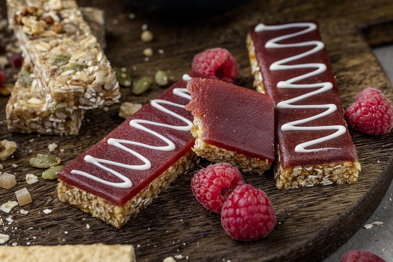 Snack bars with berry fruit topping on each of them and two white drizzle on two of them. Behind those, Behind those, snack bars with no fruit coating. Surrounding them, some seeds and raspberries.