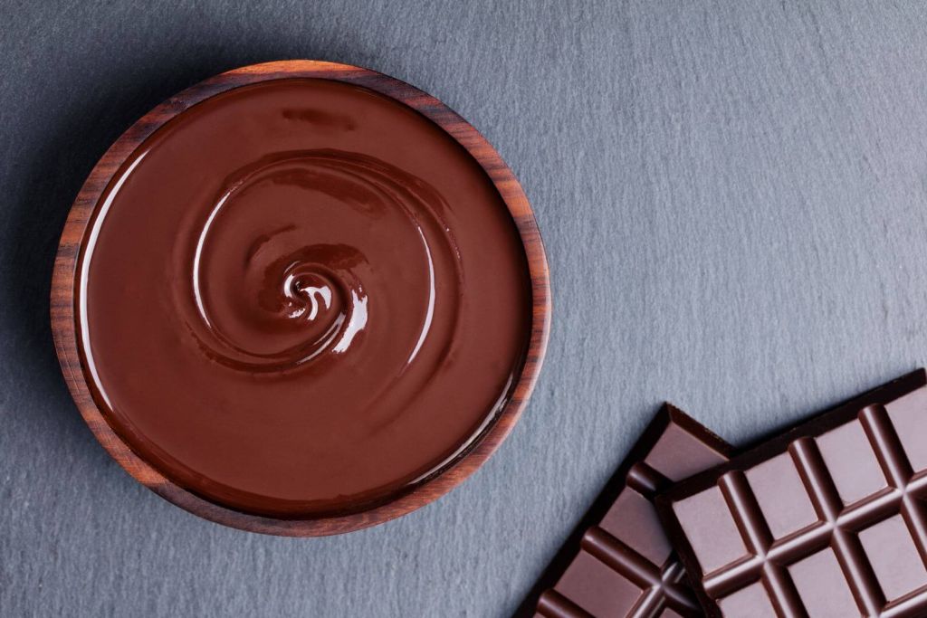 Picture taken from above of a bowl of chocolate syrup on the right and two chocolate bars on the top-left side. Sitting on a light gray counter