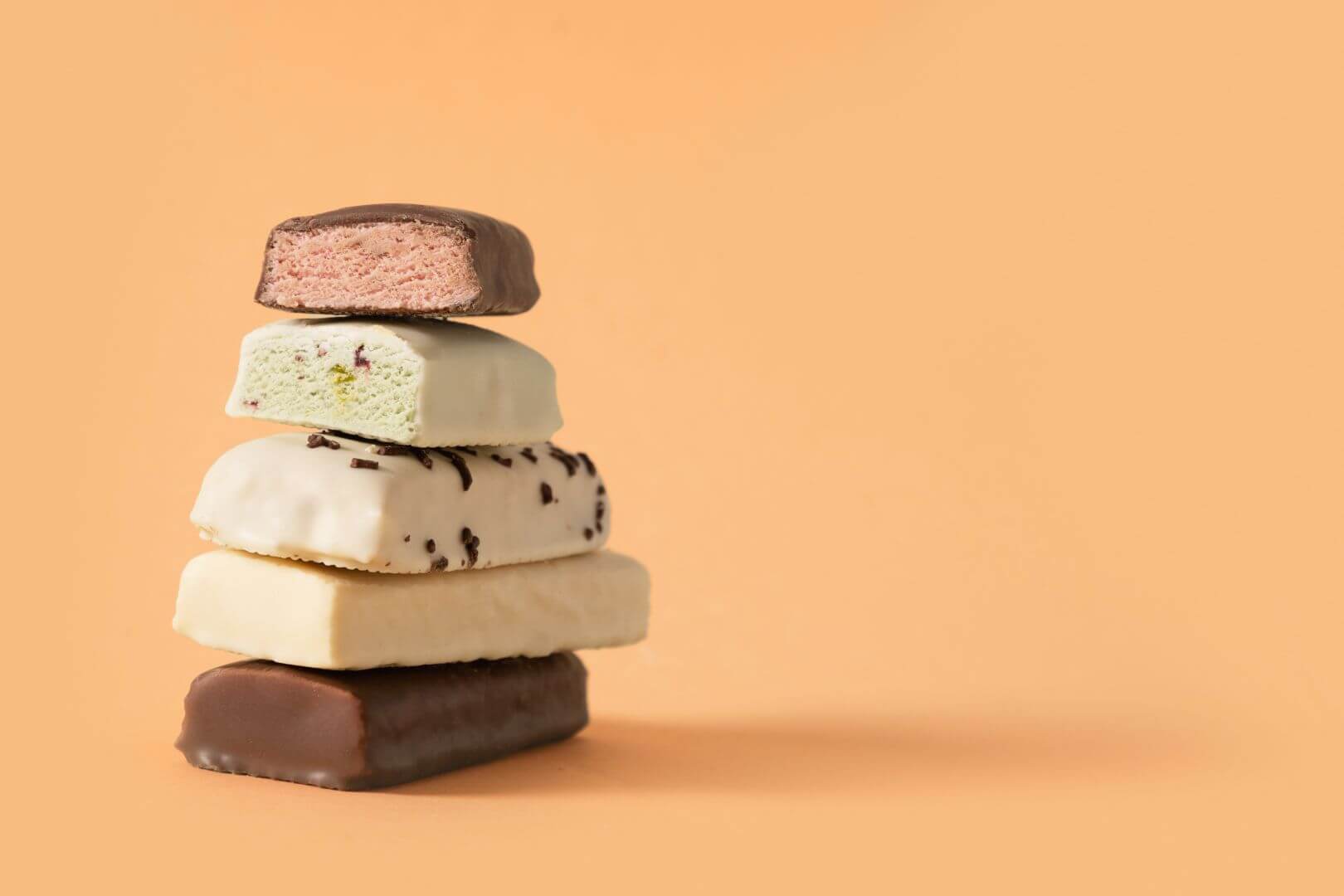 On left side of image, a stack of different chocolate, white chocolate and vanilla frosting-coated snack bars with the top bar cut in half revealing a strawberry filling. Sitting in a column on an orange background
