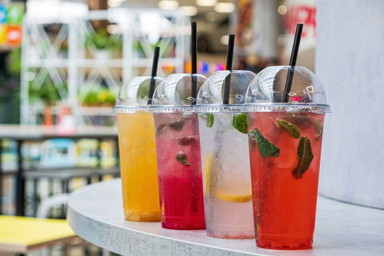 Flavored, multicolored sparkling beverages with garnishes in plastic cups with lids and straws, in a row outside
